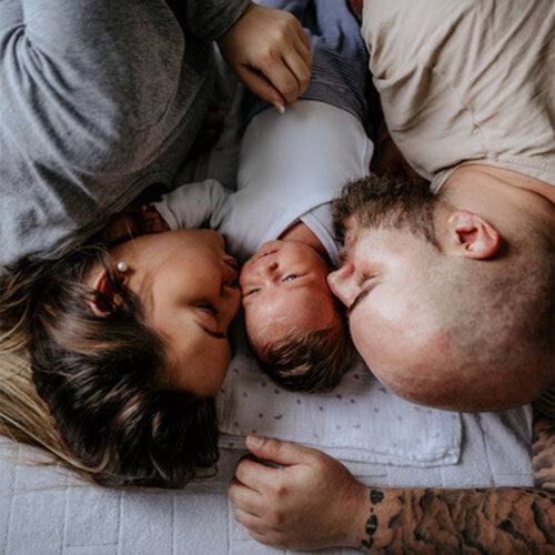 Una mamá, papá y su bebé tumbados sobre la cama besando al bebé que esta entre los dos. - solucionesbanohub.compaña - 1