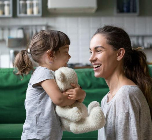 Una niña feliz abraza a su osito mientras le sonríe a su madre en la cocina de su casa - solucionesbanohub.compaña - 1