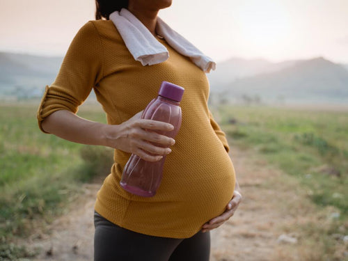 Mujer en la naturaleza con ropa de deporte y botella de agua sujetando su tripa de embarazada  - solucionesbanohub.compaña - 1