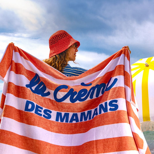 Una mujer sujetando su toalla en el playa. La toalla tiene la frase "la créme de mamans" - solucionesbanohub.compaña - 1