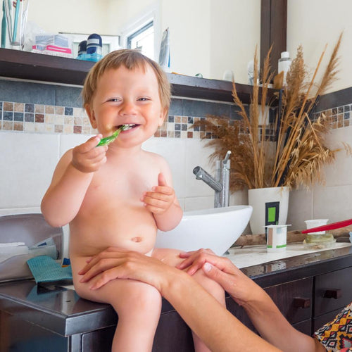 Un bebé feliz se cepilla los dientes por primera vez, sentado en el baño de su hogar y en compañía de su mamá - solucionesbanohub.compaña - 1