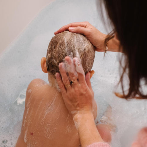 Una mamá limpia la cabeza de su bebé con champú Mustela mientras él disfruta de un baño de espuma - solucionesbanohub.compaña - 1