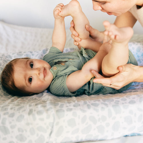 Una bebé feliz estirada en la cama mientras su mamá le ayuda a hacer ejercicios para el estreñimiento - solucionesbanohub.compaña - 1