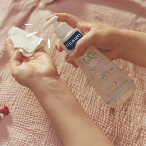 Manos aplicando el agua limpiadora de Mustela sobre un disco de algodón, con una botella con dosificador sobre un fondo rosado. - solucionesbanohub.compaña - 1