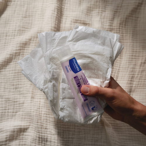 Mano sujetando la Crema bálsamo 1 2 3 de Mustela sobre unos pañales. - solucionesbanohub.compaña - 1
