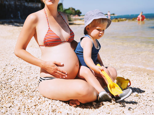 Una madre embaraza con su bebé disfruta de la playa mientras se aplica correctamente el protector solar de Mustela  - solucionesbanohub.compaña - 1