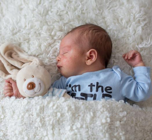Bebé durmiendo plácidamente arropado sobre una sabana blanca y con un osito de peluche. - solucionesbanohub.compaña - 1