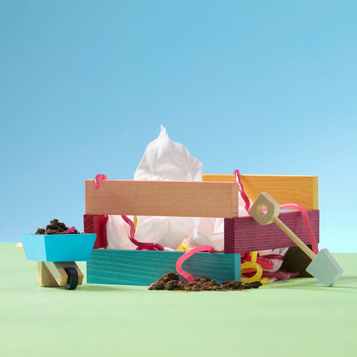 Escena colorida en miniatura con una caja de madera pintada, una servilleta blanca sobresaliendo, tiras decorativas de colores, una carretilla azul con tierra y pequeñas herramientas de jardinería sobre un fondo azul y verde. - solucionesbanohub.compaña - 1