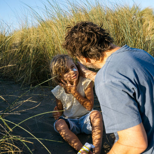 Padre junto su hijo que se esta aplicando crema solar en la cara entre dunas y hierbas altas en un día soleado. - solucionesbanohub.compaña - 1