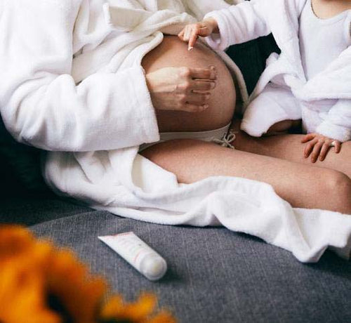 Una mamá embarazada y su hija, las dos con albornoz, después de tomar un baño y aplicándose crema Mustela en la barriga - solucionesbanohub.compaña - 1