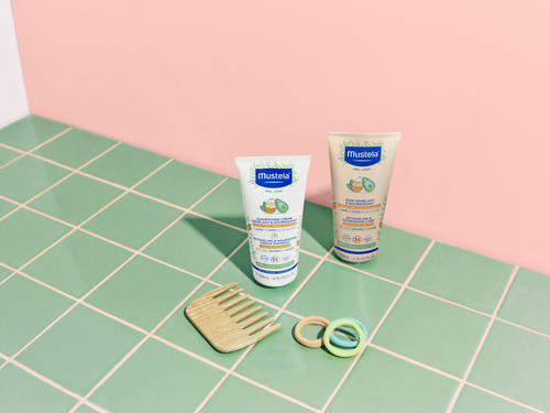 Nuevo champú y acondicionador de Mustela sobre azulejos verdes con un peine y tres coletas para el pelo de colores. - solucionesbanohub.compaña - 3