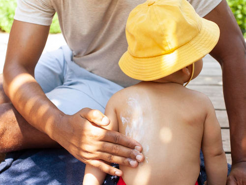 un padre aplica protector solar en la espalda de su niño que lleva un gorro. slider - solucionesbanohub.compaña - 6