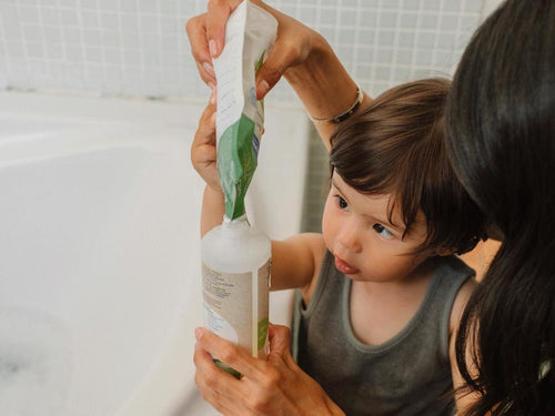 Un niño con su madre está rellenando el envase del gel-champú bio de Mustela con su eco-recarga. slider - solucionesbanohub.compaña - 5
